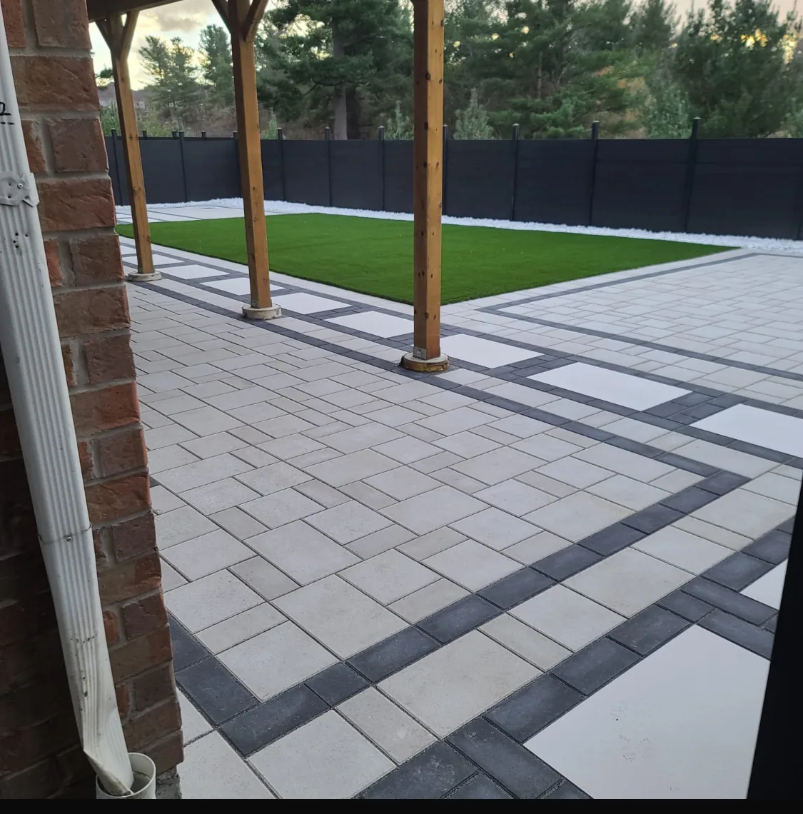Backyard after hardscaping with light pavers, dark banding borders, and an artificial turf panel
