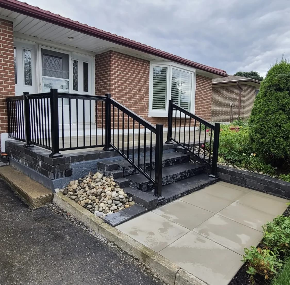 Front of house after hardscaping with block-faced steps, black railings, river rock bed, and slab walkway